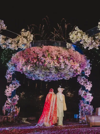 A bride and groom stand before a magnificent floral mandap, framed by an explosion of fireworks. This image captures the epic scale and romantic grandeur we strive to create for our couples.