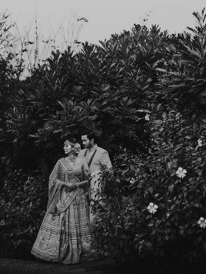 A moody black and white portrait of the couple in a garden. The deep shadows and natural framing from the foliage create a sense of intimacy and romance.