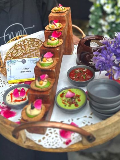 Chana Jor Garam, elevated. We serve this classic street snack on crisp mathris with fresh toppings and a duo of chutneys, all presented on an elegant wooden tray adorned with flower petals.