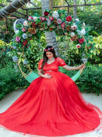 A serene mom-to-be in a flowing red gown, seated on a floral swing in a garden.
