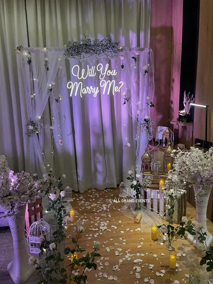 This is how we turn a simple room into a romantic proposal spot. The setup features a 'Will You Marry Me?' neon sign against a backdrop of sheer white drapes and fairy lights. We scattered white rose petals and placed flameless candles to create a warm, intimate, and absolutely magical atmosphere.