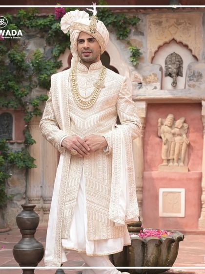 A groom in a stunning ivory sherwani, set against a backdrop of ancient sculptures. The image captures the timeless, artistic quality of the Shauhar collection.