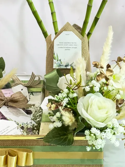 A close-up of the eco-friendly hamper, showing the beautiful arrangement of white roses, dried grasses, and a lucky bamboo plant wrapped in jute. The personalized tag adds a final thoughtful touch.