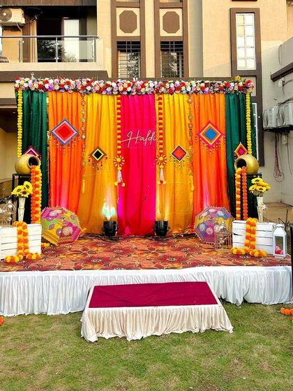 This outdoor Haldi stage setup is full of color. It combines bright drapes, marigold garlands, and decorative pots on a raised platform, perfect for a garden or lawn ceremony.