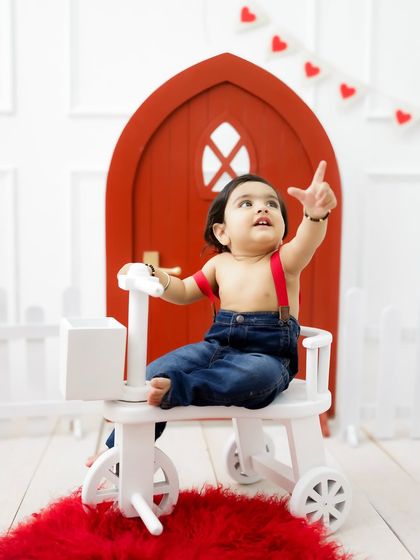 A playful moment on the tricycle, with the baby pointing up in excitement. The simple set design keeps the focus on his joyful expression.