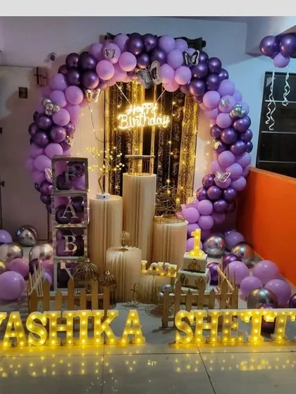 An elegant purple-themed first birthday for Yashika Shetty. This grand setup includes a balloon arch, 'BABY' blocks, pedestals for the cake, and illuminated letters spelling out the full name.