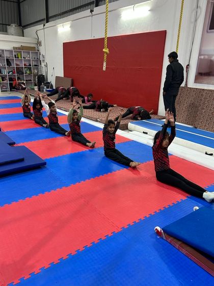 Pike stretching is essential for skills on bars and floor. Here, a group of young gymnasts works on their forward fold, lengthening their hamstrings under a coach's supervision.