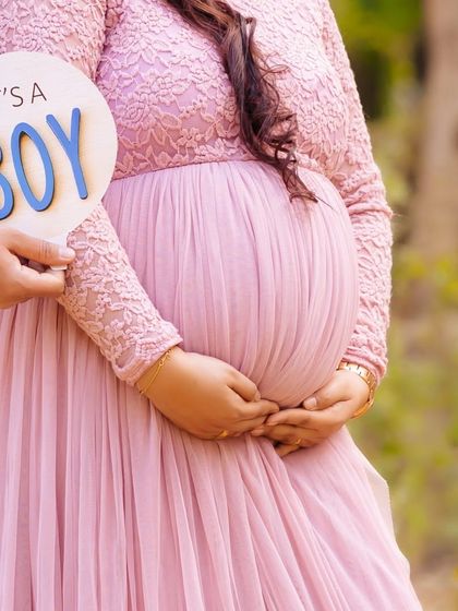 A close-up shot focusing on an "It's a BOY" sign held next to the baby bump. The soft pink of the gown and the outdoor setting create a lovely gender reveal photo.