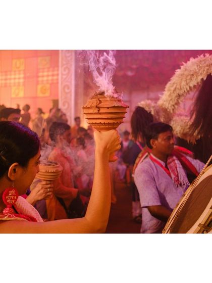A woman holds a dhunuchi aloft while a drummer plays the 'dhaak' in the background, capturing the sound and movement of the festival.