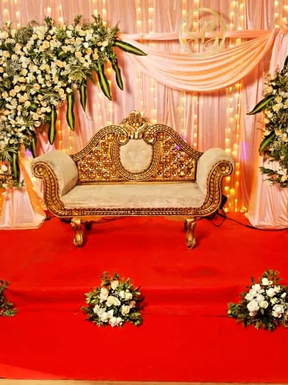 For more intimate gatherings, we design elegant stages like this one. It features a beautifully carved golden sofa, a soft draped backdrop with fairy lights, and tasteful white floral arrangements on a red carpet.