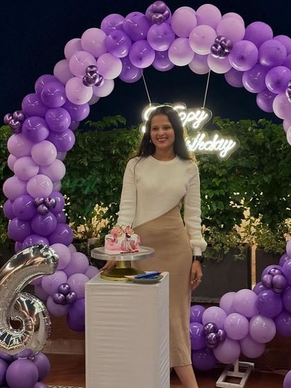 A woman posing with her cake at her 26th birthday party, in front of a purple balloon ring.