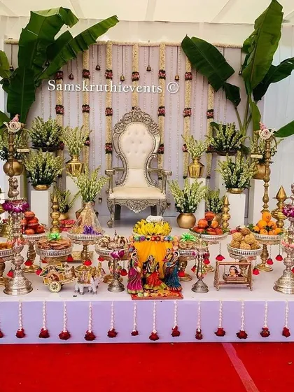 An elegant and bright Seemantha decoration featuring a white backdrop with hanging sugandharaja garlands. The stage is flanked by banana plants and a symmetrical arrangement of offerings, creating a pure and sacred ambiance.