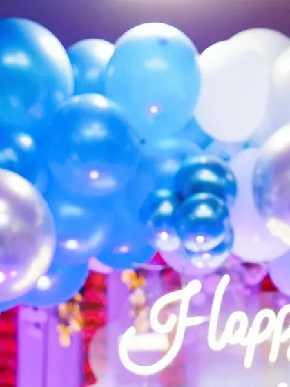 A neon "Happy" sign glows behind a cascade of blue and silver balloons, capturing the modern and fun decor of a celebration.