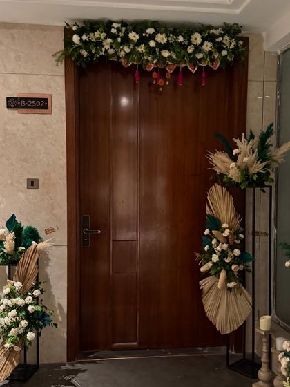 The entrance to this home was decorated for a housewarming with a beautiful floral garland over the door and two modern stands holding arrangements of white flowers, pampas grass, and dried green palms.