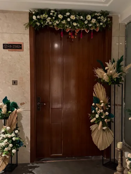 The entrance to this home was decorated for a housewarming with a beautiful floral garland over the door and two modern stands holding arrangements of white flowers, pampas grass, and dried green palms.