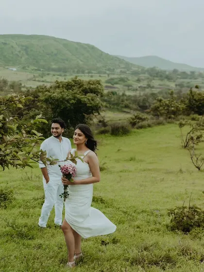 A candid pre-wedding photo of a couple in a beautiful green meadow, with the bride holding a bouquet of flowers.