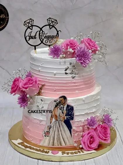 To celebrate a new beginning, we designed this romantic two-tier engagement cake with a blend of Black Forest and Blueberry flavors. It features an edible photo of the couple, soft pink ombre frosting, and fresh floral arrangements for a personal and elegant touch.