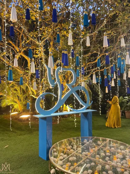 A personalized corner featuring the couple's monogram, set under a tree decorated with hanging lights and custom blue and white tassels.