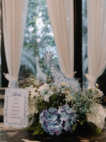 An elegant table centerpiece from a blue Dior-inspired shower. A giraffe figure stands amidst a lush arrangement of blue and white hydrangeas and baby's breath, next to a custom menu card.
