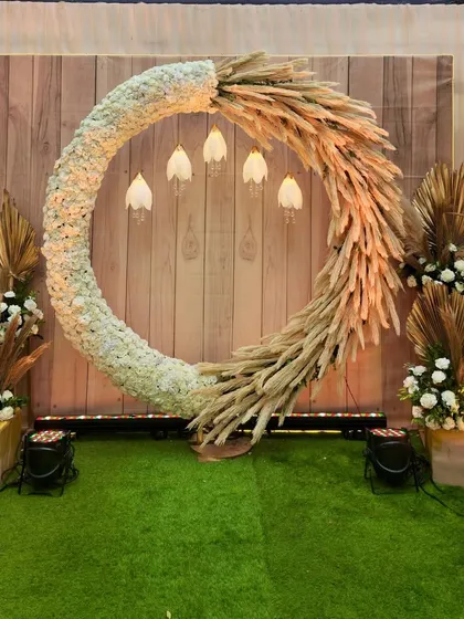 A beautiful bohemian photo booth with a large circular frame made of white flowers and pampas grass. The wooden backdrop and hanging lights add to the rustic, earthy charm.