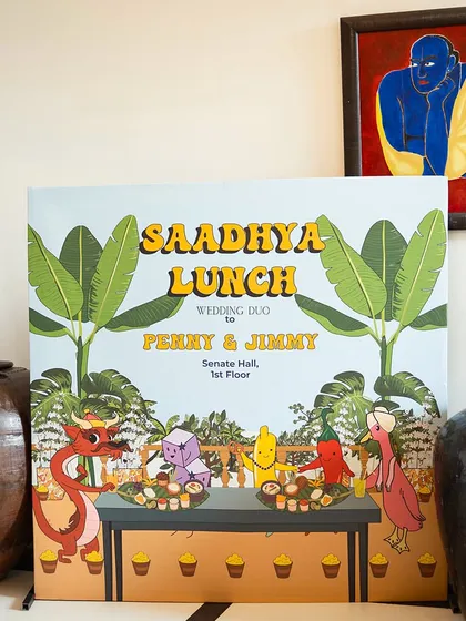 A quirky and personalized welcome sign for the 'Saadhya Lunch', featuring custom cartoon illustrations of the couple and their friends, set between traditional pottery.