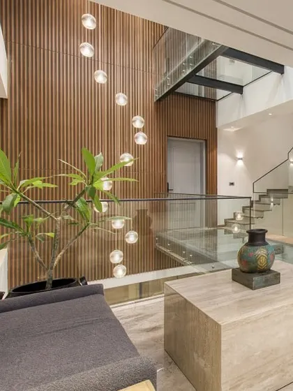 The triple-height atrium in the Kedias Residence, featuring a glass bridge that connects the upper floors. The cascading pendant lights and wood-paneled wall add warmth and drama to the space.