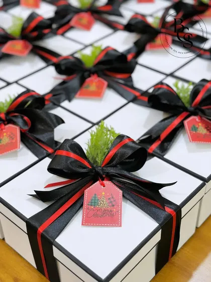 A large order of Christmas gifts in crisp white boxes. Each gift is wrapped with a combination of black and red ribbons and finished with a festive tag and a small green sprig, creating a chic and coordinated holiday look.
