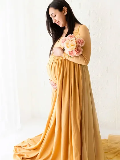 A full-length portrait of a mother-to-be in a long yellow gown, holding a bouquet of flowers. The soft, natural light from the window enhances the gentle mood of this boho session.