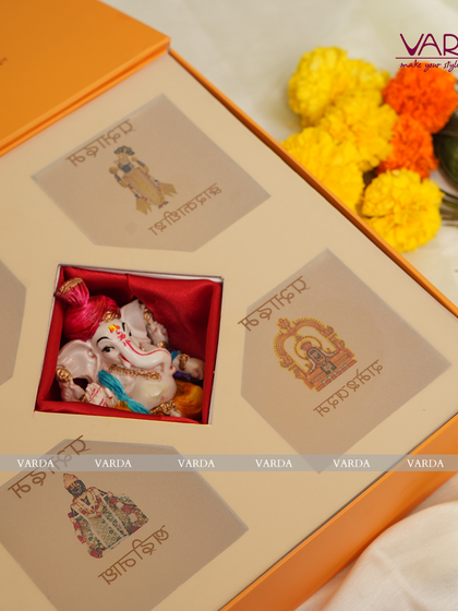 This bright orange gift box is designed for a sacred ceremony, featuring a miniature Ganesha idol nestled in its own compartment. The surrounding spaces can be filled with sweets or other small gifts, each covered with a card depicting a different deity.