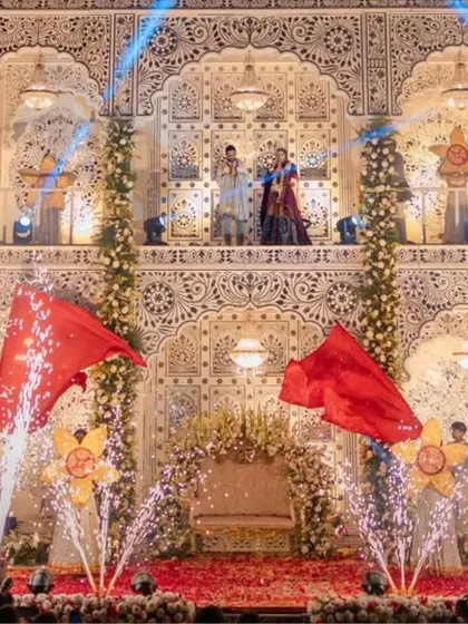 A close-up of a royal varmala ceremony. The couple stands on an elevated platform against an ornate backdrop, with cold pyros and performers adding to the grandeur of the moment.