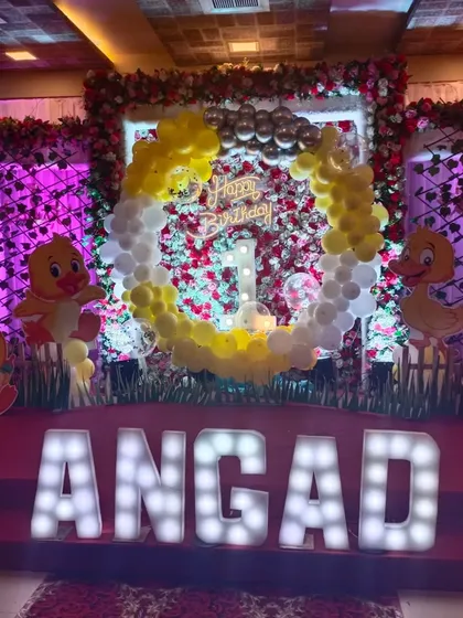 A cute duck-themed first birthday decoration. The backdrop is a mix of flowers and balloons, with large marquee letters for the name 'Angad' making a great centerpiece.