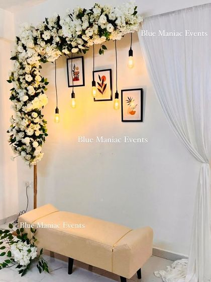 A minimalist and modern engagement setup. This design features an L-shaped floral stand with white flowers, paired with hanging Edison bulbs, framed pictures, and a simple bench for a chic and personal touch.