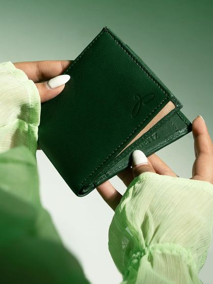 A close-up of hands with white nails opening a green leather wallet. The soft green fabric of the sleeves complements the wallet perfectly.