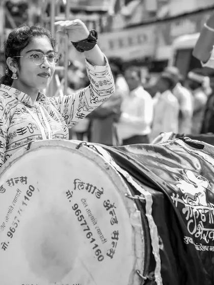 A black and white portrait of a female dhol player with glasses, her hand raised.