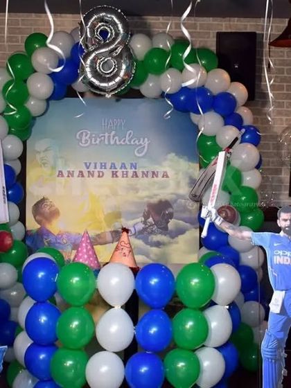 A cricket-themed birthday party for an 8-year-old fan. The decor includes a custom backdrop and cutouts of famous cricketers, set within a green, blue, and white balloon arch.