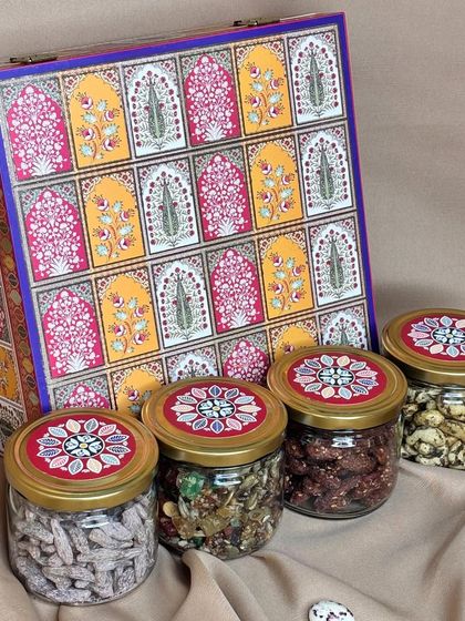 This hamper showcases a beautiful box with a traditional 'jharokha' design, paired with four glass jars filled with assorted dry fruits and nuts. The lids are coordinated with the box design for a complete, elegant look.