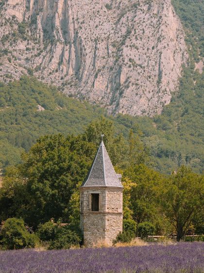 A stone tower stands at the edge of a lavender field, with a dramatic mountain cliff in the background. This image captures the rustic charm of the Provence region.