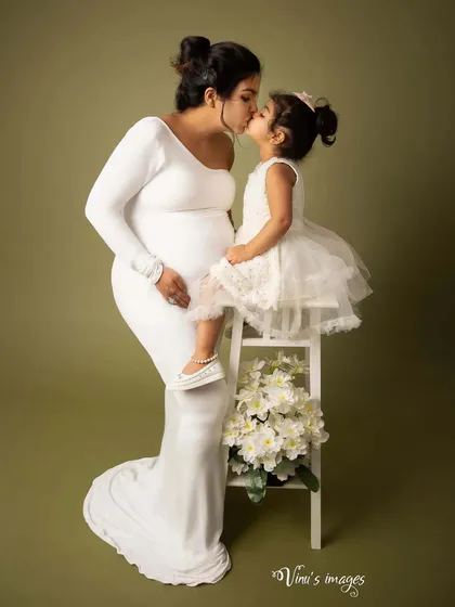 A pure and tender moment between a mother and her daughter. Dressed in white, the mother kisses her daughter, who is perched on a stool, creating a beautiful and heartfelt maternity portrait.