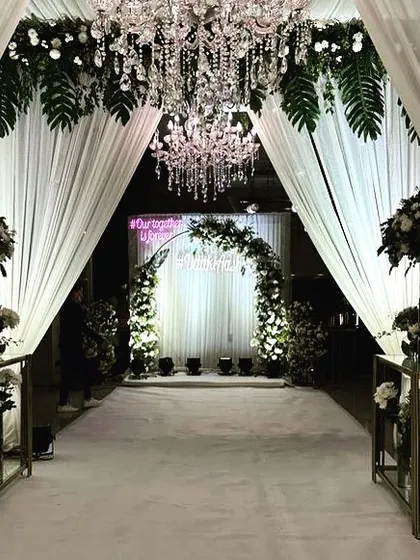 A dreamy entrance for a Roka ceremony, featuring a white carpeted walkway, elegant drapes, and a ceiling adorned with greenery and crystal chandeliers, leading to a floral photo-op arch.