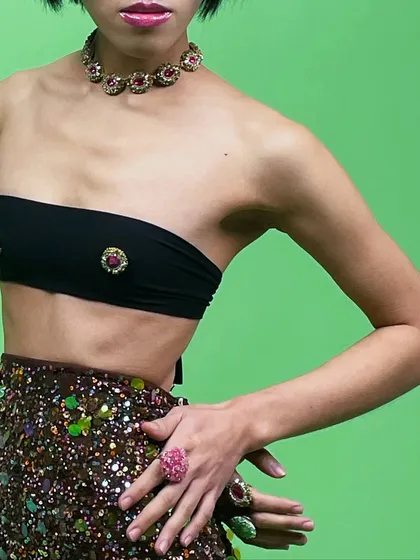 A close-up on a jewelry set against a vibrant green screen. The styling is bold and playful, with a focus on the colorful, oversized rings and choker.