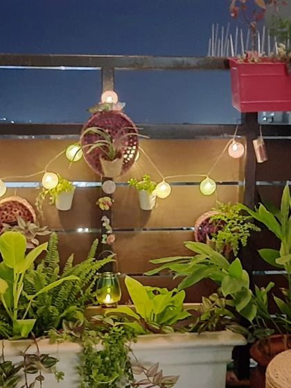 A cozy balcony corner decorated with string lights for a festive feel. The mix of ferns and other tropical plants in various planters creates a warm and inviting atmosphere, perfect for a quiet evening.