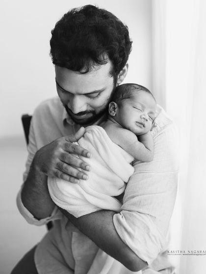 A father holds his sleeping newborn, swaddled in white. This black and white portrait, taken near a window, is full of soft light and quiet emotion.