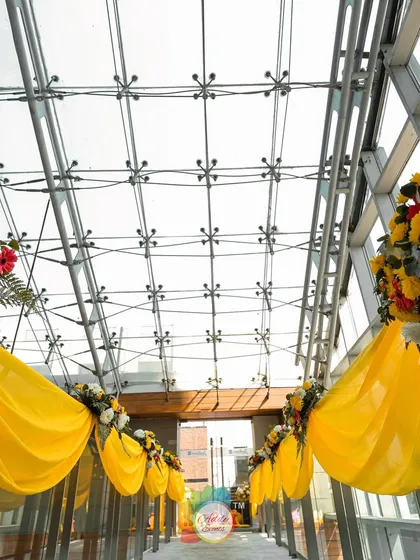 A sunlit glass-covered walkway decorated with yellow drapes and floral bouquets for the Haldi ceremony.
