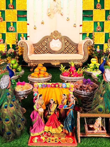 This decoration for a Seemantham features a traditional yellow and green checkered backdrop. The setup includes a beautiful sofa, peacock figures, and a detailed doll arrangement depicting the baby shower ceremony.