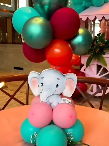 A cute elephant-themed table centerpiece. A small elephant cutout sits atop a colorful arrangement of pink, red, and teal balloons.