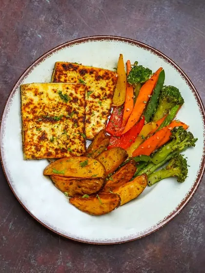 A vegetarian platter with seasoned grilled paneer, potato wedges, and sautéed vegetables. The dark background makes the colors of the vegetables and the char on the paneer stand out.