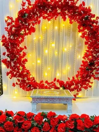 This modern Ganpati decoration features a bold circular ring completely covered in vibrant red artificial flowers. Set against a curtain of fairy lights, this design provides a contemporary and striking focal point for your celebration.