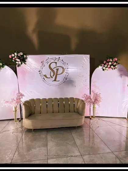 A chic and modern reception stage with a minimalist aesthetic, featuring three white arched panels, a custom gold monogram, and a stylish beige sofa, accented with simple floral bouquets on tall stands.