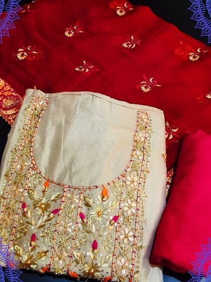 An off-white unstitched suit set with heavy gota patti and threadwork in shades of pink and orange. It is paired with a bright red Banarasi dupatta for a rich, festive look.