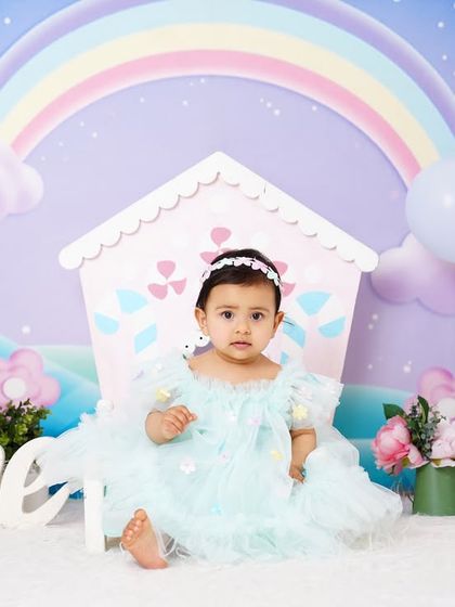 A full view of a whimsical first birthday setup, with a baby girl in a pastel blue dress surrounded by props and a rainbow backdrop.
